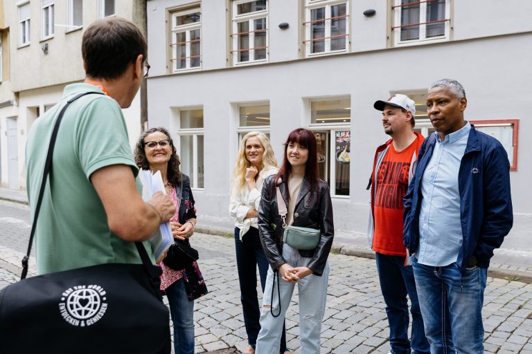 Ein Reiseleiter führt eine bunt gemischte Gruppe von fünf Erwachsenen auf einer historischen Tour und spricht zu ihnen, während sie auf einer Kopfsteinpflasterstraße im Herzen der Koblenzer Altstadt stehen.