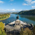 Luftaufnahme des Deutschen Ecks in der Koblenzer Altstadt, wo sich zwei Flüsse treffen, umgeben von Bäumen und Fahnen - ein Highlight auf jeder historischen Tour.