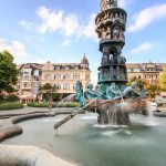 Der kunstvolle Brunnen mit Statuen und Wasserstrahlen auf einem von Bäumen und historischen Gebäuden umgebenen Stadtplatz fängt den Charme der Koblenzer Altstadt in der Nähe des berühmten Deutschen Ecks ein - ein unverzichtbarer Halt auf jeder Historischen Tour.