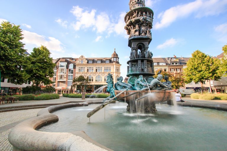 Der kunstvolle Brunnen mit Statuen und Wasserstrahlen auf einem von Bäumen und historischen Gebäuden umgebenen Stadtplatz fängt den Charme der Koblenzer Altstadt in der Nähe des berühmten Deutschen Ecks ein - ein unverzichtbarer Halt auf jeder Historischen Tour.