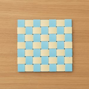 Eine quadratische Matte mit blauem und cremefarbenem Schachbrettmuster, perfekt als Bastelunterlage für geflochtene Osterkörbchen, die auf einer hellen Holzfläche ausgestellt werden.