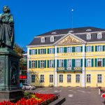 Statue vor einem gelben Gebäude mit grünen Fensterläden und roten Blumen an einem sonnigen Tag in der Bonner Innenstadt, ein perfekter Ausgangspunkt für eine kulinarische Stadtführung.