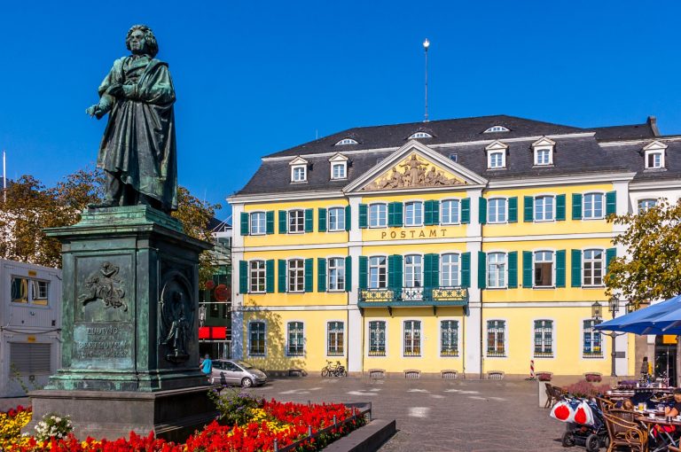 Statue vor einem gelben Gebäude mit grünen Fensterläden und roten Blumen an einem sonnigen Tag in der Bonner Innenstadt, ein perfekter Ausgangspunkt für eine kulinarische Stadtführung.