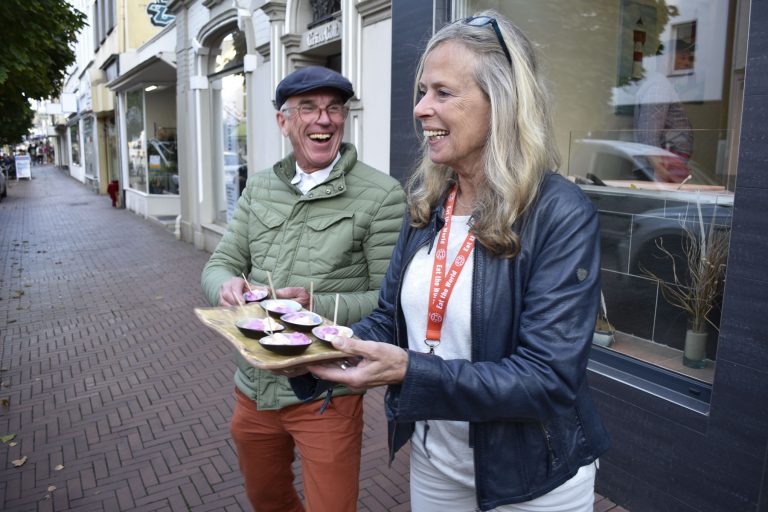 Zwei lächelnde Menschen servieren kleine Desserts auf einem Tablett, während sie auf einem Bürgersteig in Bonn stehen und dieser kulinarischen Stadtführung durch die Innenstadt Charme verleihen.