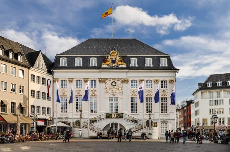 Historisches weißes Gebäude mit verzierter Fassade und Fahnen in der Bonner Innenstadt, umgeben von Menschen, die den belebten Platz und den blauen Himmel genießen - perfekt für eine kulinarische Stadtführung.