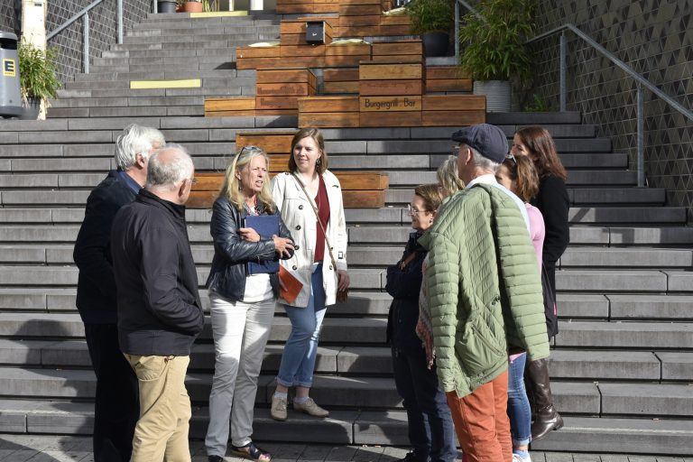 Eine Gruppe von Menschen steht auf einer Freitreppe in der Nähe von Holzkübeln und einem Gebäude und unterhält sich. Vielleicht halten sie während einer kulinarischen Stadtführung Bonn inne, um die lebendige Atmosphäre der Bonner Innenstadt zu genießen.