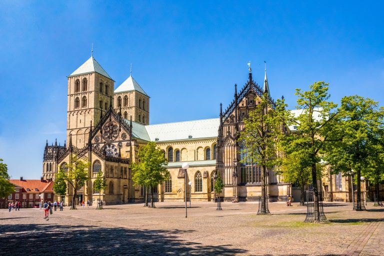 Der historische Münsterdom mit seinen Zwillingstürmen und seiner gotischen Architektur steht stolz in der Altstadt, umgeben von Bäumen und einem großen Platz.