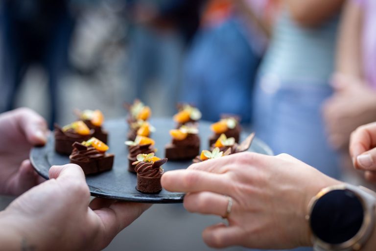 Hände, die bei einem Treffen in Münsters Altstadt ein Tablett mit kleinen, verzierten Schokoladendesserts mit Orangenbelag anbieten - die perfekten Leckerbissen für die nächste kulinarische Stadtführung.