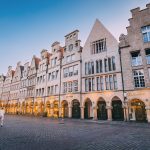 In der kopfsteingepflasterten Altstadt von Münster spazieren die Menschen bei Sonnenuntergang an historischen Giebelhäusern und Geschäften vorbei.