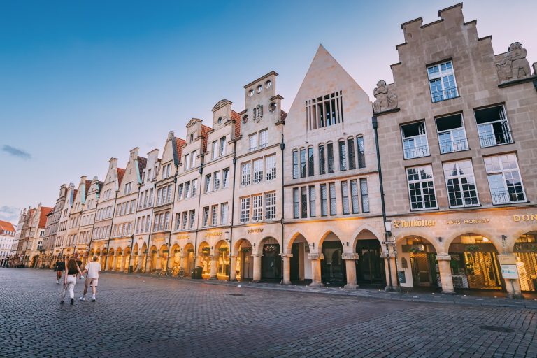 In der kopfsteingepflasterten Altstadt von Münster spazieren die Menschen bei Sonnenuntergang an historischen Giebelhäusern und Geschäften vorbei.