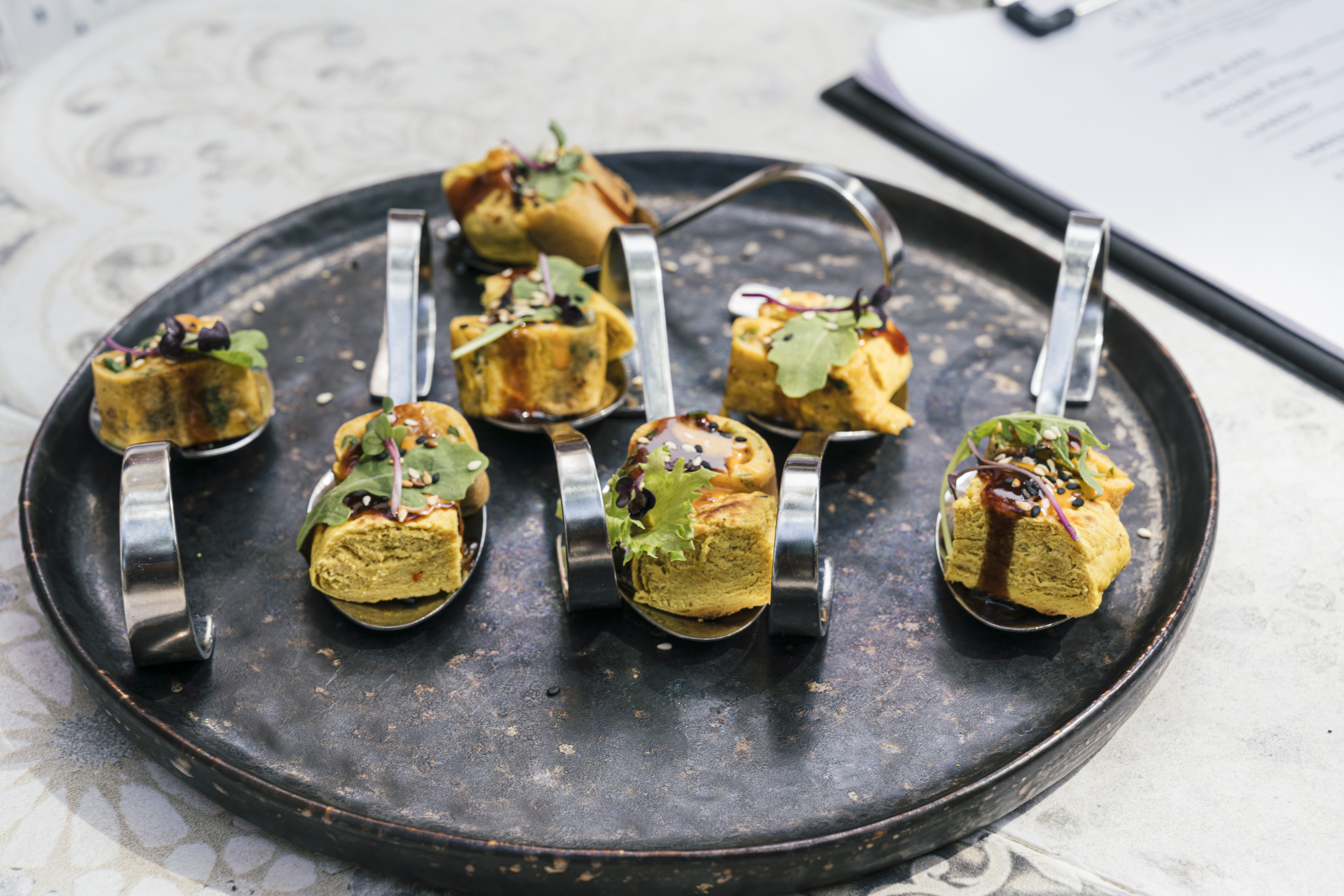 Acht Gourmet-Häppchen mit Grünzeug und Soße, serviert auf Metalllöffeln auf einem runden schwarzen Tablett - perfekt für einen Spaziergang zu Leipzigs Sehenswürdigkeiten.