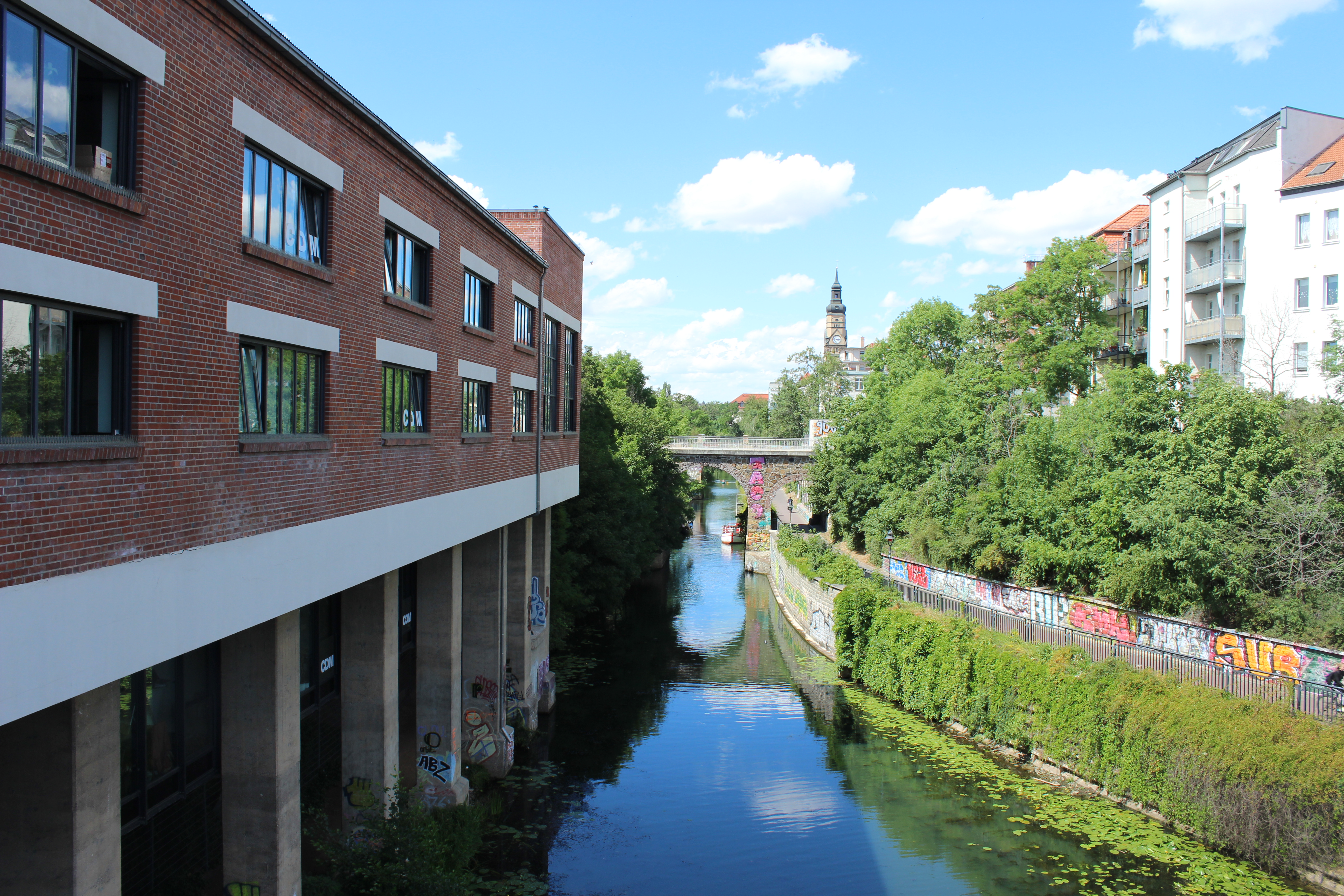 Ein Kanal verläuft zwischen weißen Backsteinbauten in Leipzig, mit Grün und Graffiti, unter einem teilweise bewölkten blauen Himmel - perfekt für einen Spaziergang, um lokale Sehenswürdigkeiten zu erkunden.