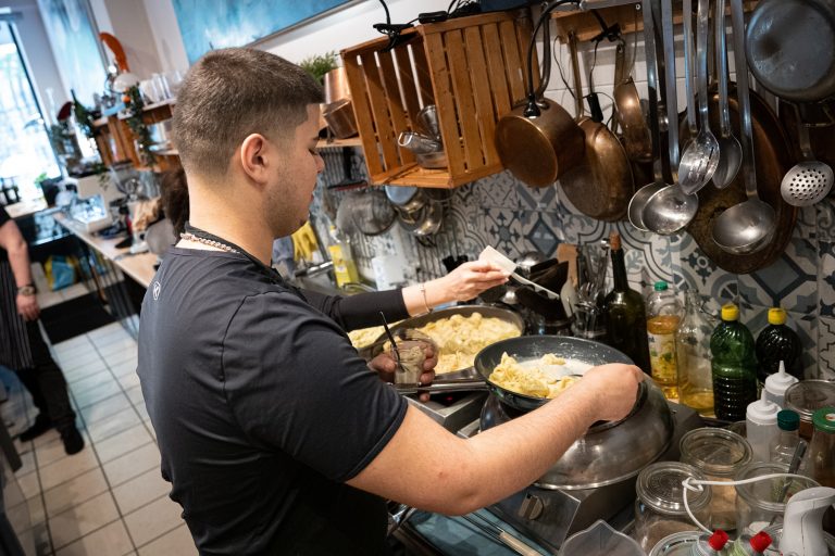Ein Mann, der in einer geschäftigen Küche Pasta zubereitet und das Essen mit einer Zange auf Tellern anrichtet, umgeben von verschiedenen Küchenutensilien und Zutaten – perfekt für jeden Entdecker und Genießer, der seinen Tag in Köln kulinarisch bereichern möchte.