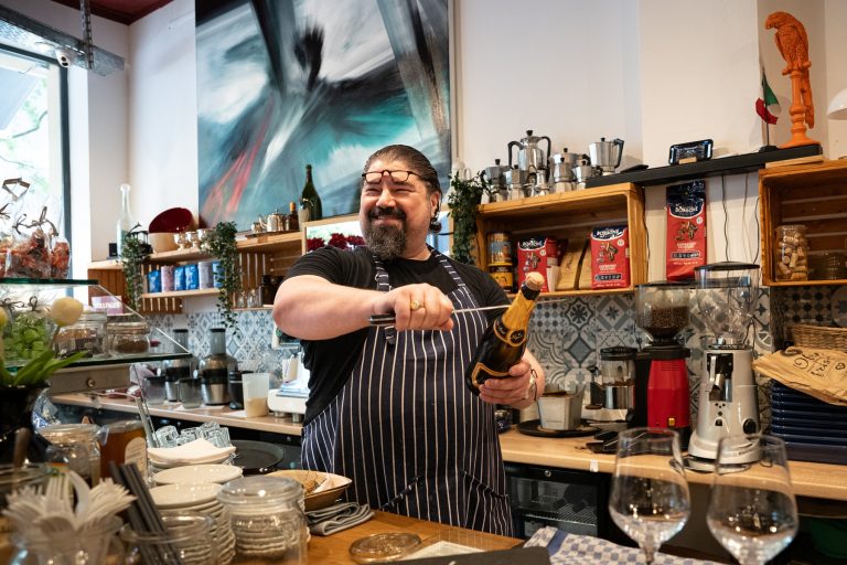 Ein lächelnder Mann in einer Schürze öffnet hinter der geschäftigen, farbenfrohen Theke eines Cafés eine Flasche Champagner und heißt jeden Entdecker und Genießer herzlich willkommen, um ihnen den Tag in Köln zu versüßen.