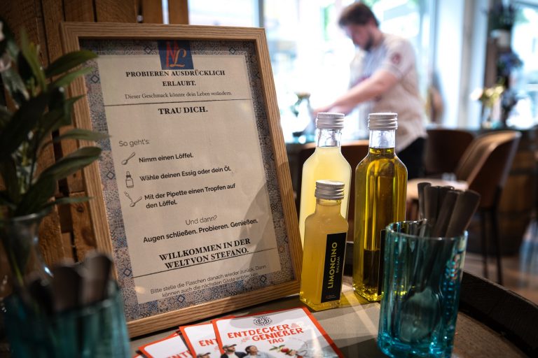 Ein gerahmtes Schild mit Verkostungshinweisen, Flaschen mit Limoncino und Öl sowie Broschüren auf einer Café-Theke – perfekt für alle, die den „Entdecker Genießer Tag Köln“ genießen.