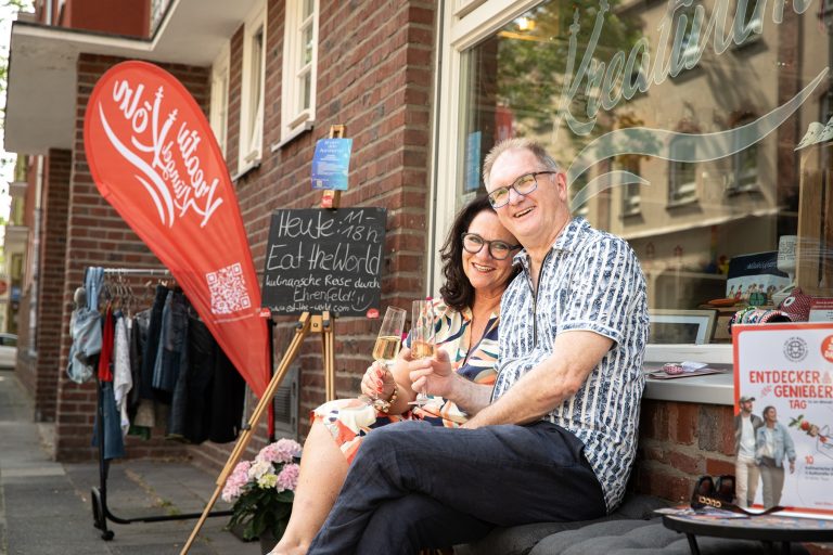 Ein lächelndes Paar sitzt vor einem Geschäft und genießt an einem sonnigen „Entdecker Genießer Tag“ in Köln ein Getränk, während in der Nähe Veranstaltungsschilder und Dekorationen zu sehen sind.
