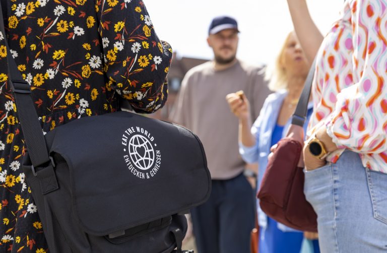 Menschen in farbenfroher Kleidung, einer mit einer schwarzen Tasche mit der Aufschrift "Eat the World Entdecken & Genießen", genießen eine Kulinarische Stadtführung Nürnbergs durch die Altstadt.