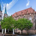 In der Nürnberger Altstadt erhebt sich eine große gotische Kirche mit hohen Türmen und Rundbogenfenstern, davor Bäume und darüber ein blauer Himmel - ein ideales Wahrzeichen für eine St. Sebald-Tour oder eine Kulinarische Stadtführung.
