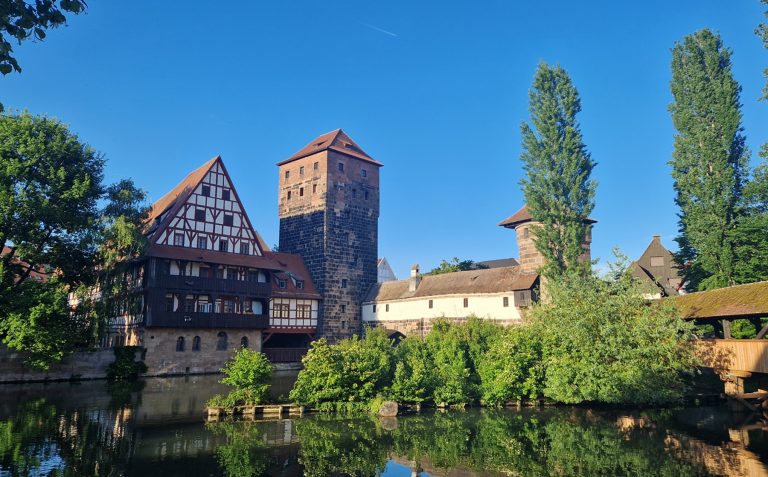 Historische Fachwerkhäuser und ein Turm am Fluss unter strahlend blauem Himmel, umgeben von Bäumen, machen den Charme der Nürnberger Altstadt bei St. Sebald aus.