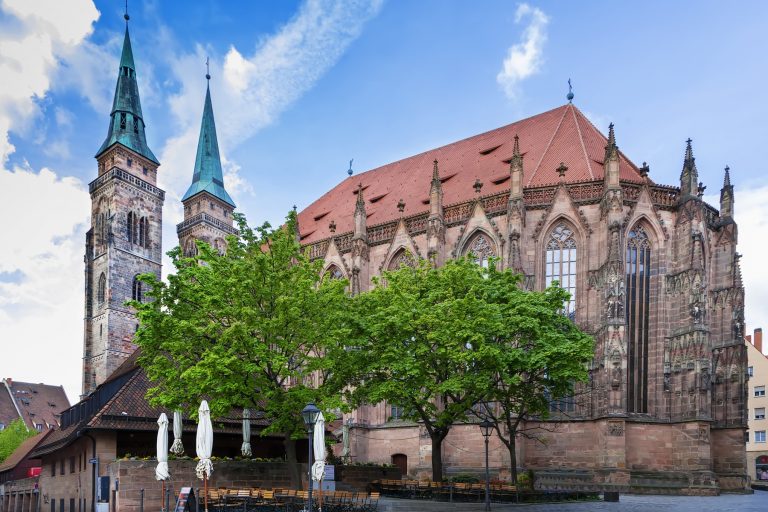 In der Nürnberger Altstadt erhebt sich eine große gotische Kirche mit hohen Türmen und Rundbogenfenstern, davor Bäume und darüber ein blauer Himmel - ein ideales Wahrzeichen für eine St. Sebald-Tour oder eine Kulinarische Stadtführung.