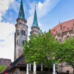 Hinter belaubten Bäumen und geschlossenen Schirmen erheben sich die Zwillingskirchtürme eines Straßencafés in der Altstadt St. Sebald, eine charmante Station auf jeder kulinarischen Stadtführung Nürnbergs unter blauem Himmel.