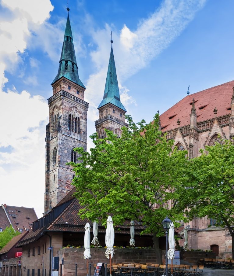 Hinter belaubten Bäumen und geschlossenen Schirmen erheben sich die Zwillingskirchtürme eines Straßencafés in der Altstadt St. Sebald, eine charmante Station auf jeder kulinarischen Stadtführung Nürnbergs unter blauem Himmel.