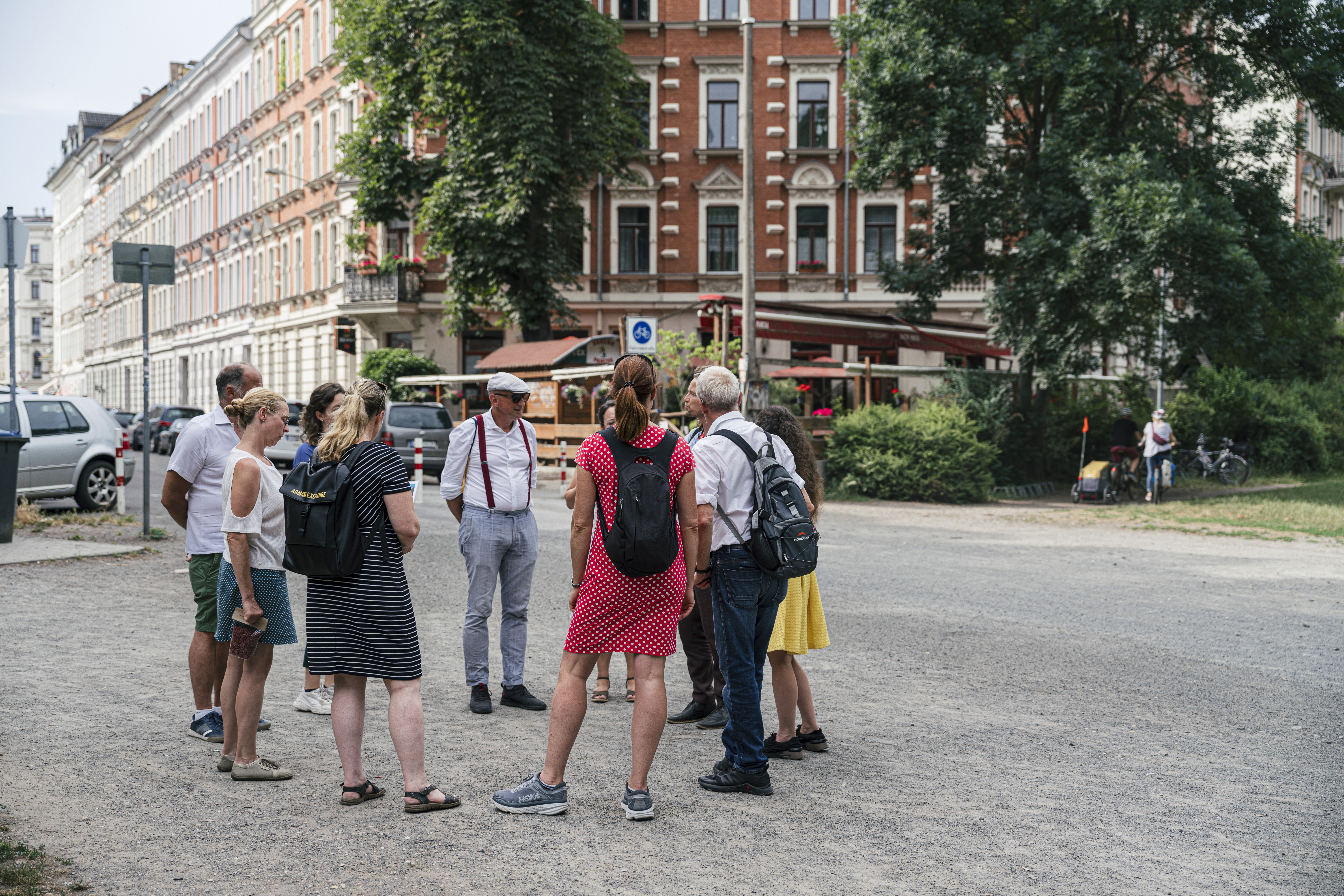 Eine Gruppe von Menschen unterhält sich an einem sonnigen Tag bei einem Spaziergang in Leipzig, in der Nähe von Bäumen und historischen Gebäuden, die zu den Sehenswürdigkeiten der Stadt gehören.