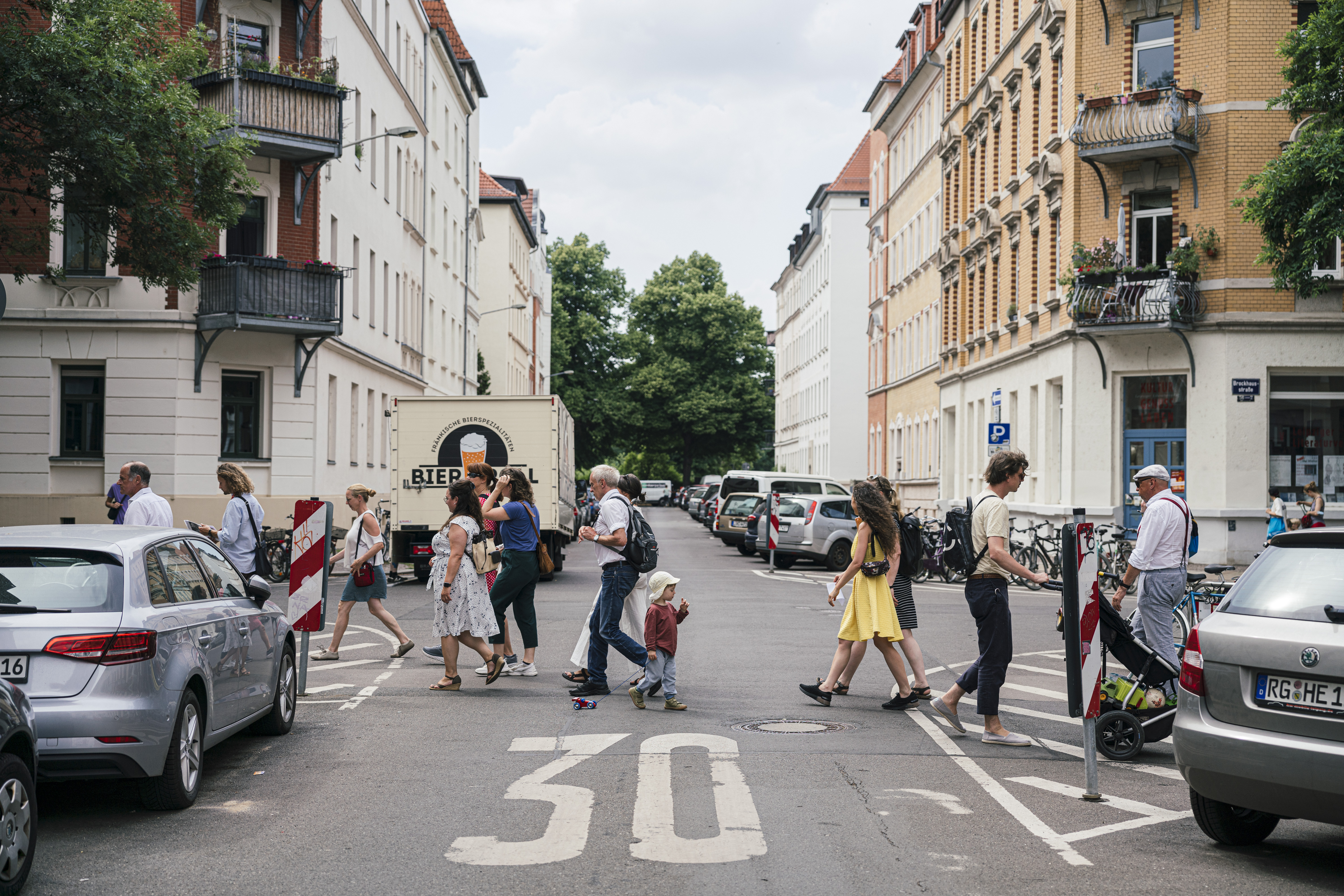 Menschen auf einem Tagesausflug Leipzig überqueren eine Stadtstraße an einem Zebrastreifen, umgeben von geparkten Autos und Wohnhäusern.