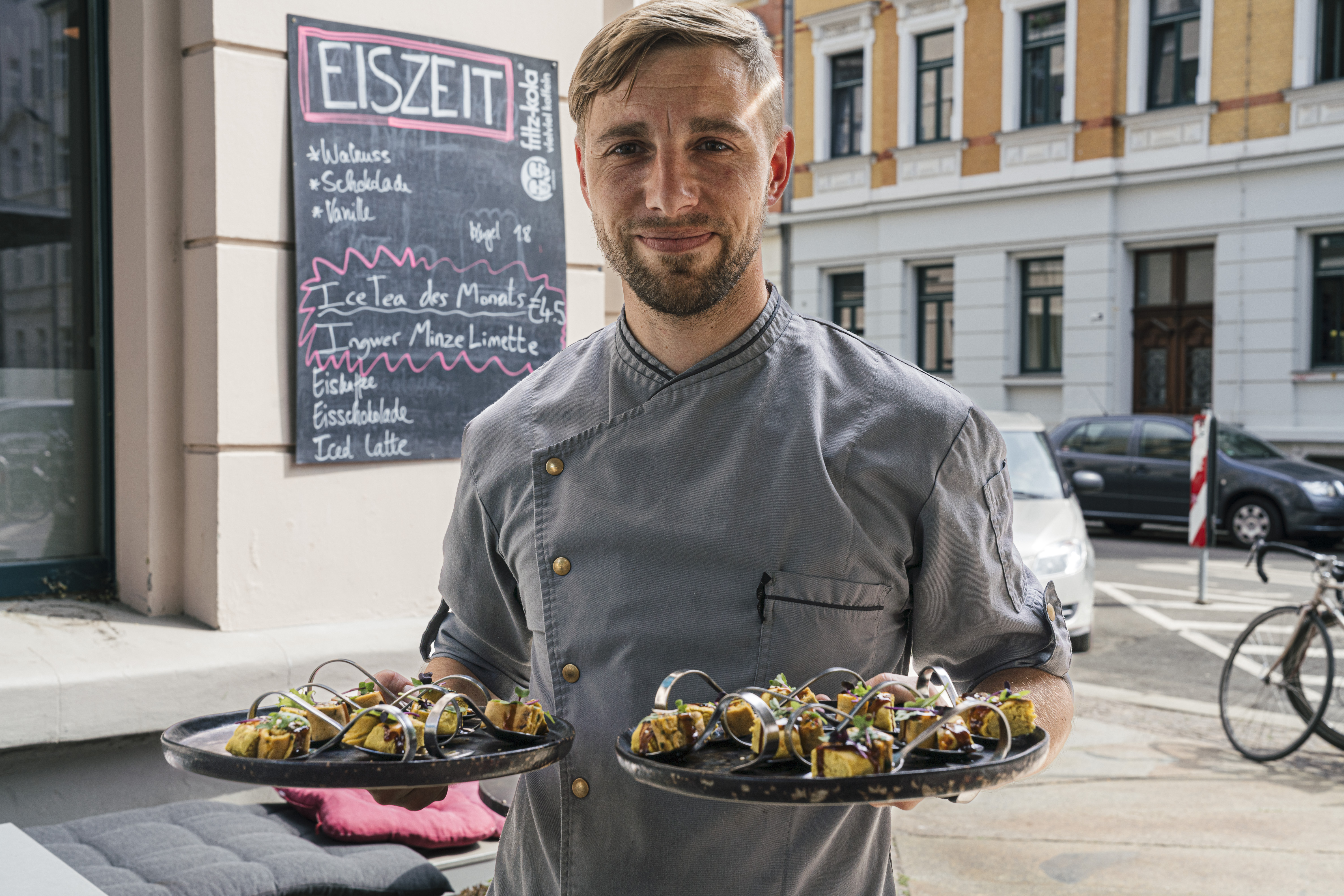 Ein Koch in grauer Uniform hält vor einem Leipziger Restaurant zwei Teller mit Vorspeisen in der Hand, hinter ihm die Speisekarte auf einer Kreidetafel - perfekt für einen leckeren Zwischenstopp auf dem Tagesausflug.