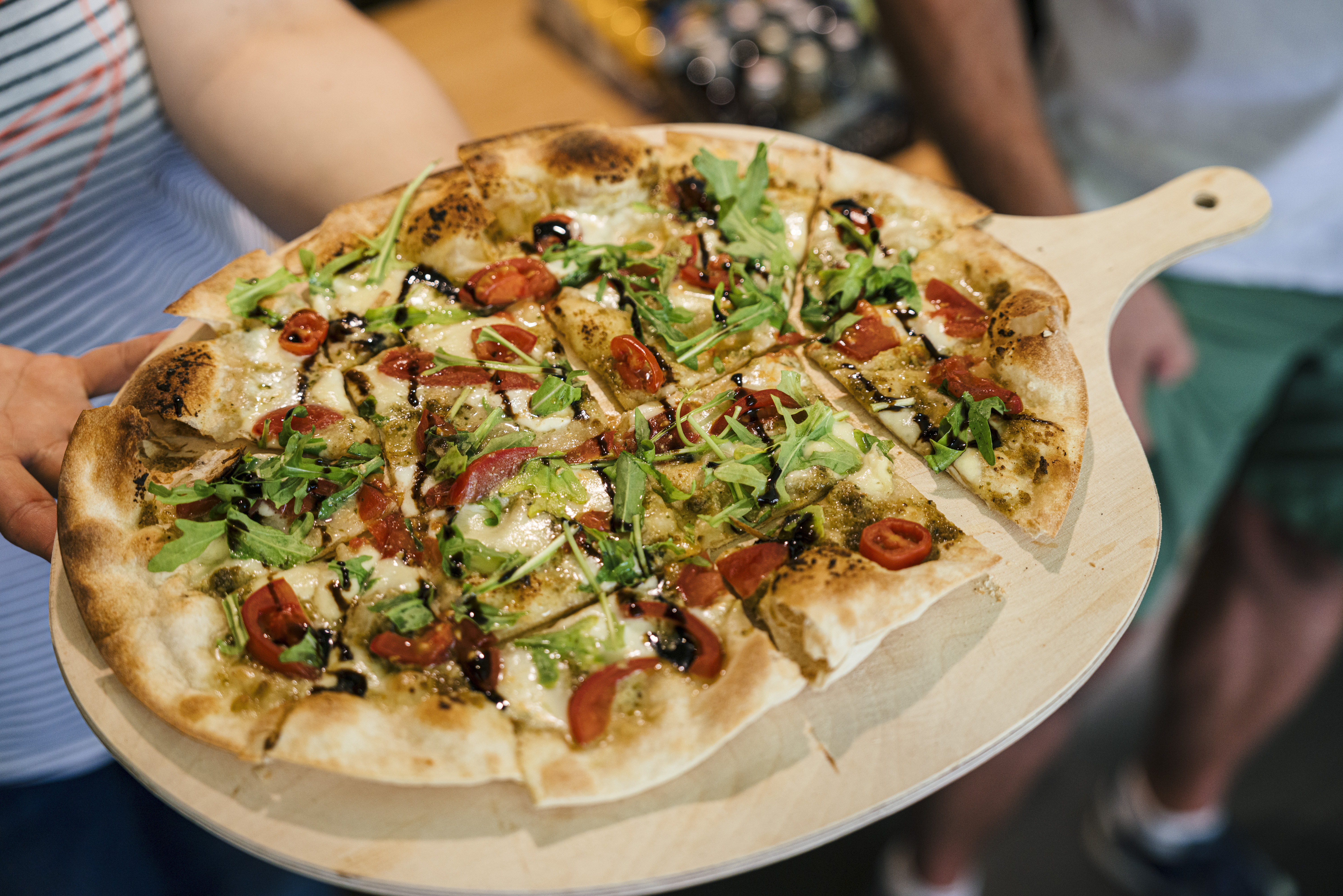 Eine Person hält eine frisch gebackene Pizza mit Rucola, Kirschtomaten und Balsamico-Glasur in der Hand - perfekt für eine leckere Pause während Ihres Tagesausflugs Leipzig.