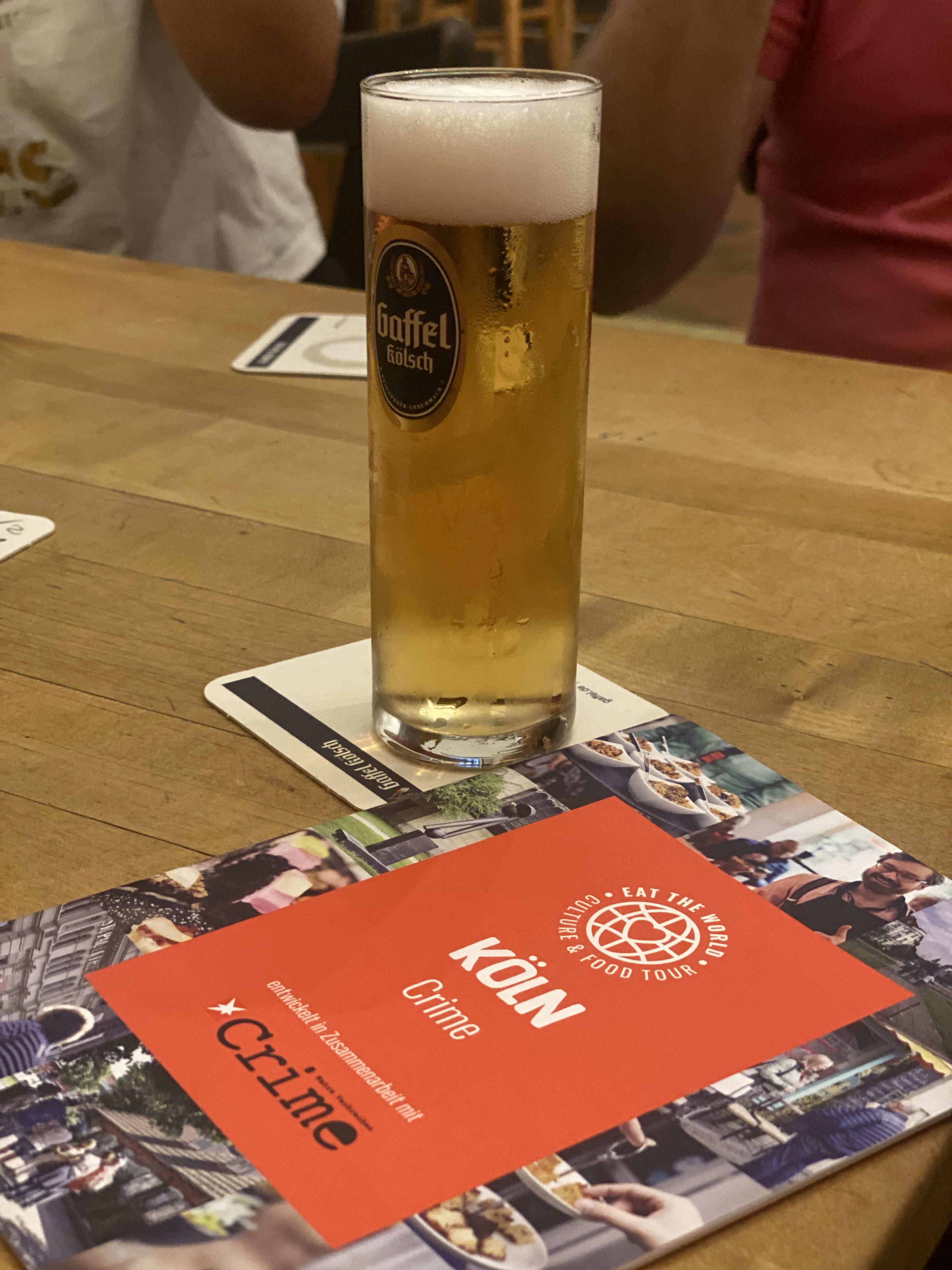 Ein Glas Gaffel Kölsch auf einem Tisch neben einer roten Köln-Food-Tour-Broschüre, perfekt für die Planung Ihres nächsten Tagesausflugs in der Stadt.