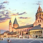 Menschen, die auf einem Platz in der Nähe des historischen Mainzer Doms in der Südlichen Altstadt unter einem farbenfrohen Sonnenuntergangshimmel spazieren gehen und die lebendige Atmosphäre dieser für ihre kulinarische Stadtführung bekannten Stadt genießen.