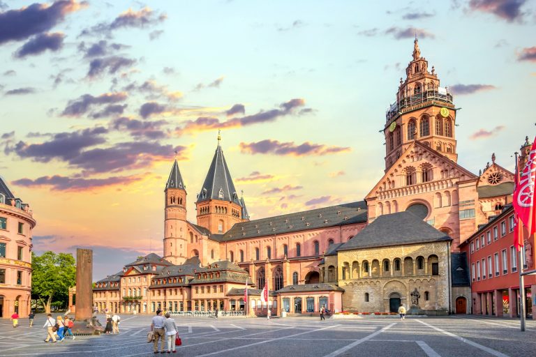 Menschen, die auf einem Platz in der Nähe des historischen Mainzer Doms in der Südlichen Altstadt unter einem farbenfrohen Sonnenuntergangshimmel spazieren gehen und die lebendige Atmosphäre dieser für ihre kulinarische Stadtführung bekannten Stadt genießen.