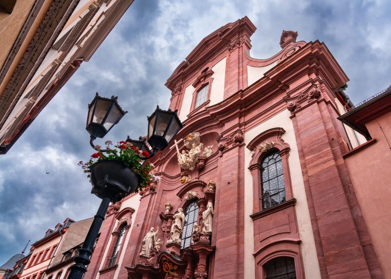 Barocke Kirchenfassade mit Statuen und Straßenlaternen unter einem dramatischen, wolkenverhangenen Himmel in der Südlichen Altstadt von Mainz, perfekt zu bewundern während einer Kulinarischen Stadtführung.