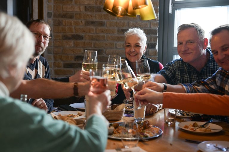 Sechs Erwachsene sitzen um einen Tisch, lächeln und stoßen mit Weingläsern auf ein Essen in einem gemütlichen Restaurant in der Südlichen Altstadt von Mainz an und genießen die Aromen ihrer kulinarischen Stadtführung.