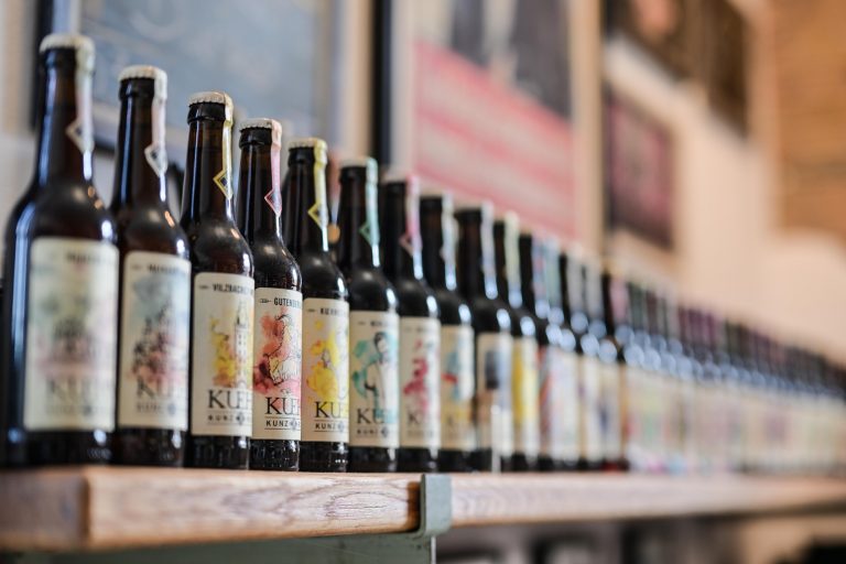 Eine Reihe von Craft-Bier-Flaschen mit farbenfrohen Etiketten, aufgereiht in einem Holzregal in einer Brauerei oder Bar in der Südlichen Altstadt von Mainz - perfekt, um sie bei einer kulinarischen Stadtführung zu entdecken.