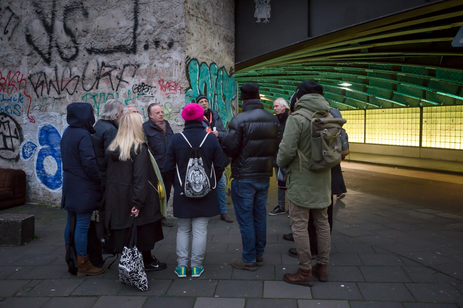 Eine Gruppe von Menschen versammelt sich unter einer mit Graffiti beschmierten Brücke auf einer Stadtstraße und entdeckt einen der Geheimtipps Köln bei ihrem Spaziergang durch das schönste Viertel Kölns.