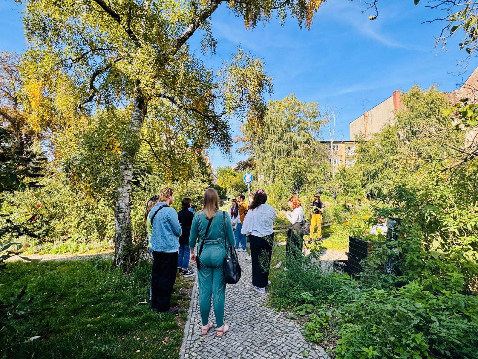 Menschen, die ihren Urlaub zu Hause genießen, stehen auf einem sonnigen Gartenweg und unterhalten sich, umgeben von üppigen Bäumen und viel Grün direkt vor ihrer Haustür.