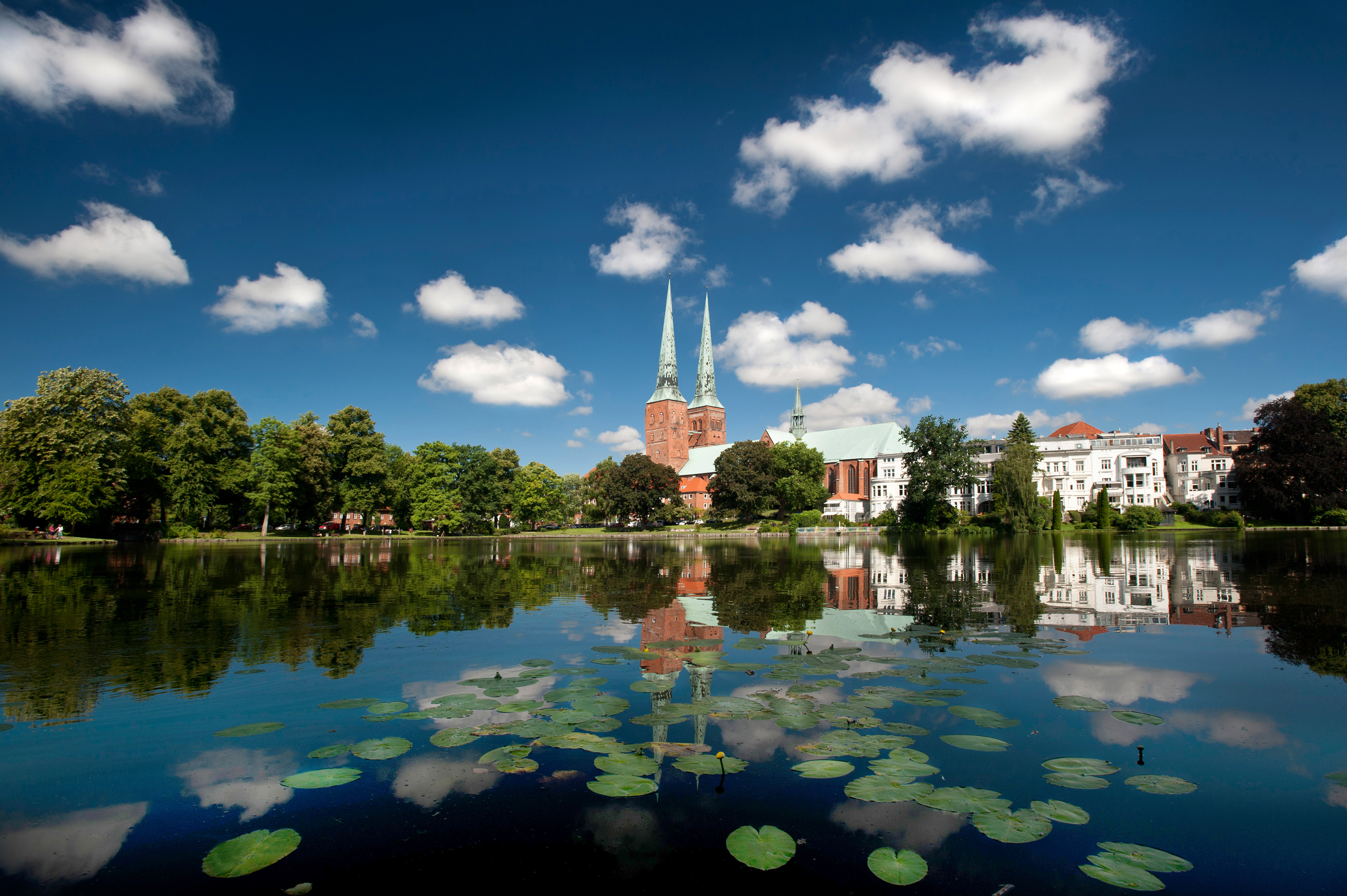 Ein ruhiger See mit Seerosenblättern spiegelt eine Kirche mit zwei Türmen und die nahegelegenen weißen Gebäude unter blauem Himmel wider – perfekt für einen „Staycation“ oder einen Urlaub vor der eigenen Haustür.