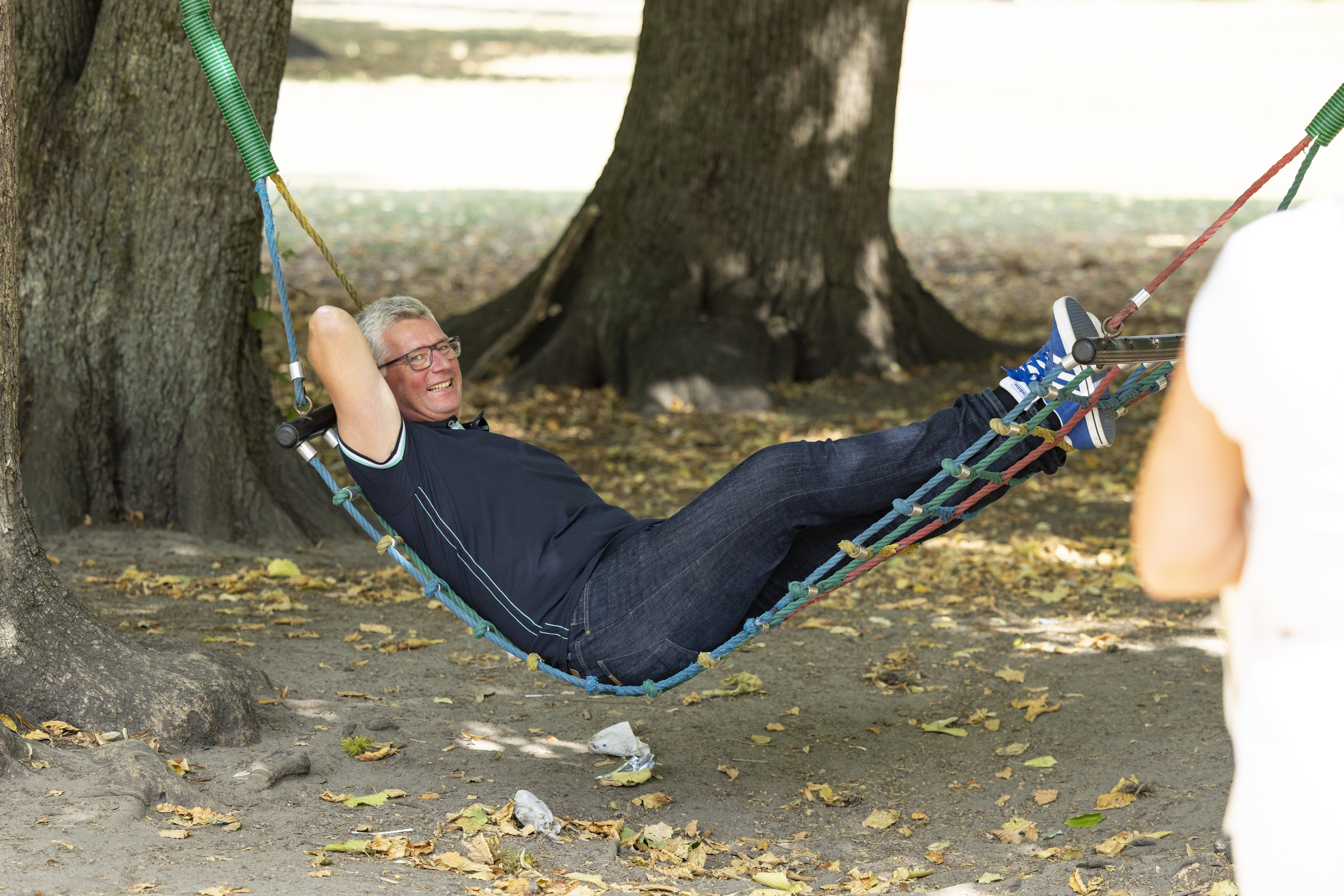 Eine lächelnde Person entspannt sich in einer Hängematte, die zwischen zwei Bäumen in einem Park gespannt ist, während Herbstblätter auf dem Boden liegen – und genießt einen gemütlichen „Staycation“-Urlaub direkt vor der eigenen Haustür.