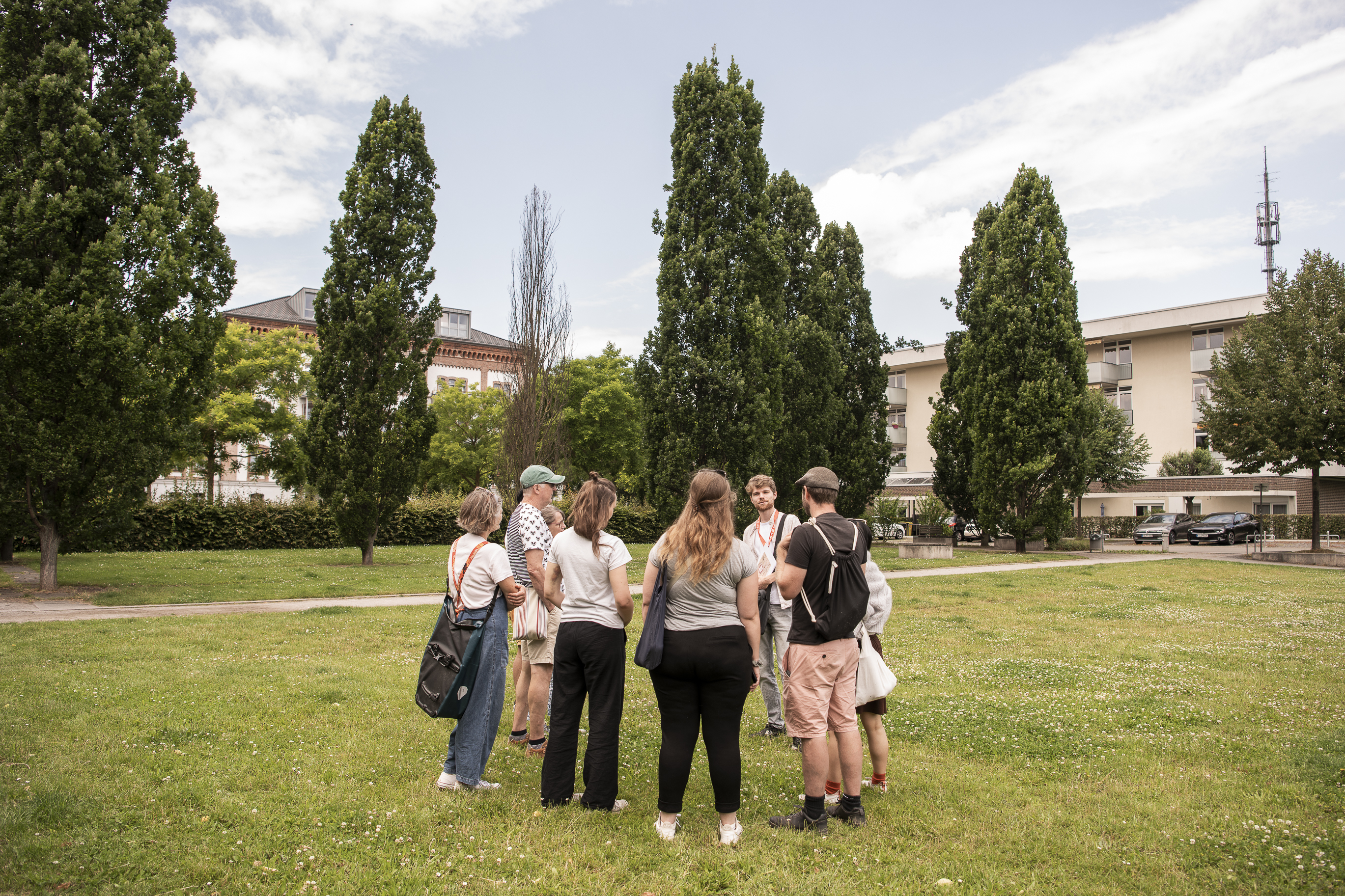 Eine Gruppe von Menschen steht gemeinsam auf einer Wiese in einem Park, der von hohen Bäumen und Gebäuden umgeben ist, und tauscht Ideen und Tipps für ihren nächsten Kurzurlaub aus.