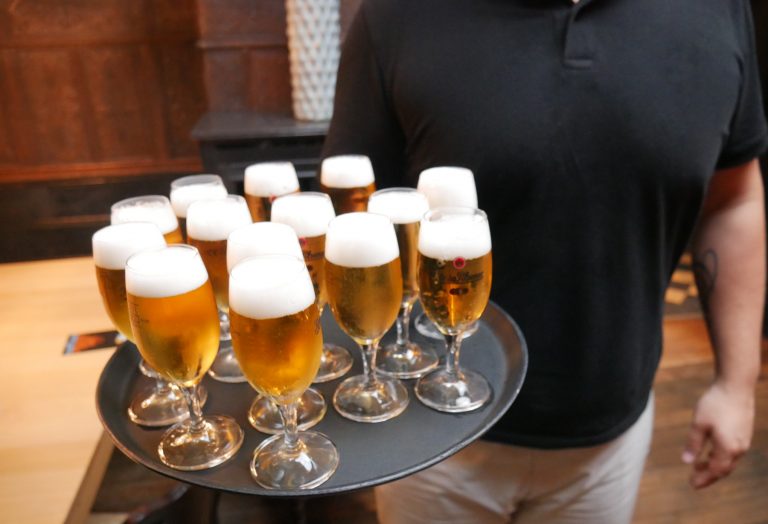 Eine Person, die während einer kulinarischen Stadtführung im Dortmunder Kreuzviertel ein Tablett mit zwölf Gläsern Bier vom Fass mit Schaum trägt.