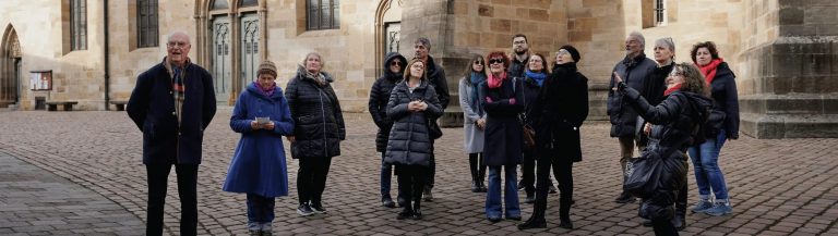 Eine Gruppe von Menschen in Winterkleidung steht gemeinsam vor einem Steingebäude, blickt nach oben und lächelt – vielleicht inspiriert von Geschichten über spannende Tätigkeiten als Reiseleiter oder von den vielen Stellenangeboten für Reiseleiter in der Region.