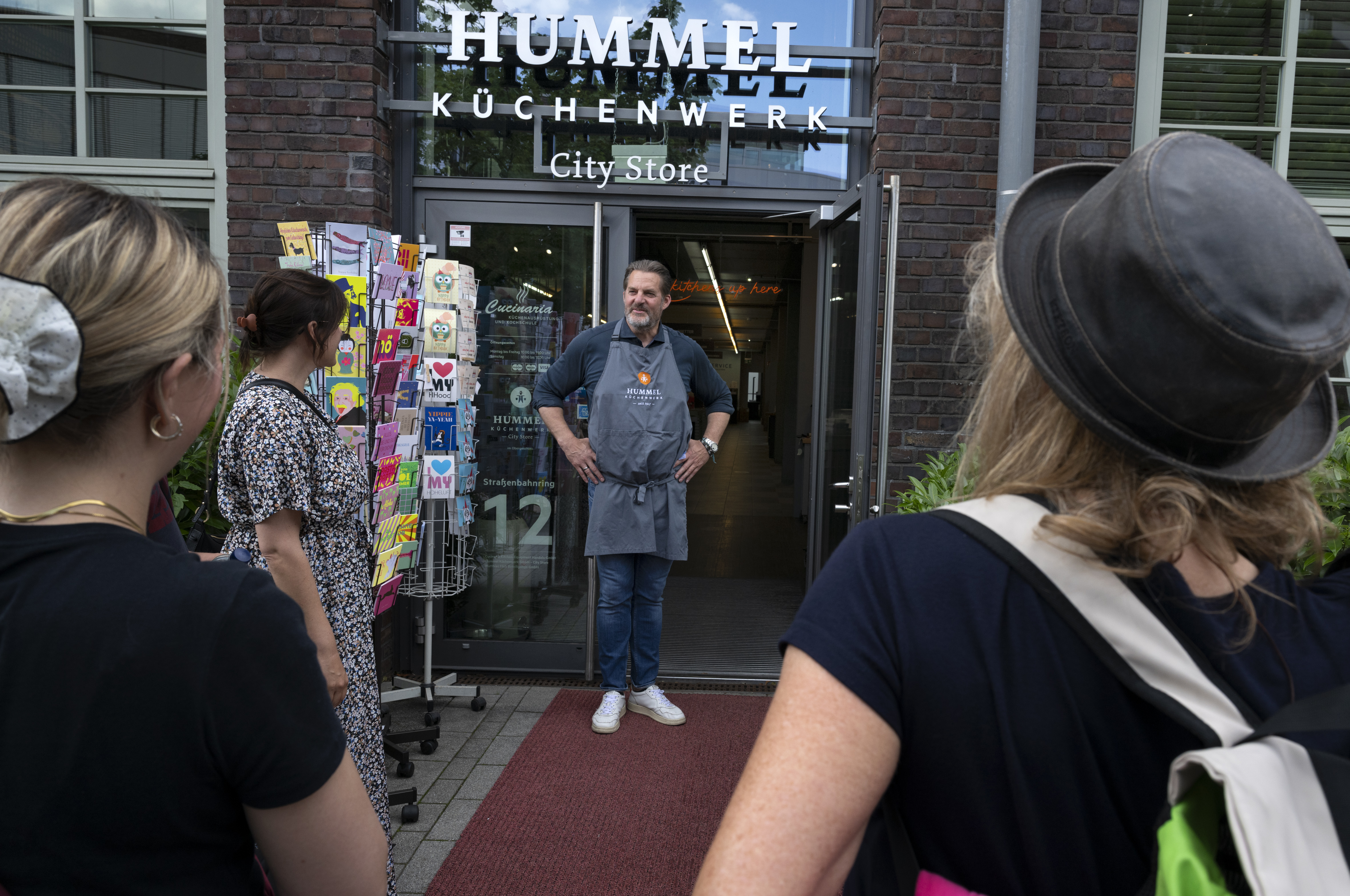 Ein Mann in einer Schürze steht am Eingang des Geschäfts „Hummel Küchenwerk“ im charmanten Viertel Hoheluft in Hamburg, während draußen Menschen zuschauen.