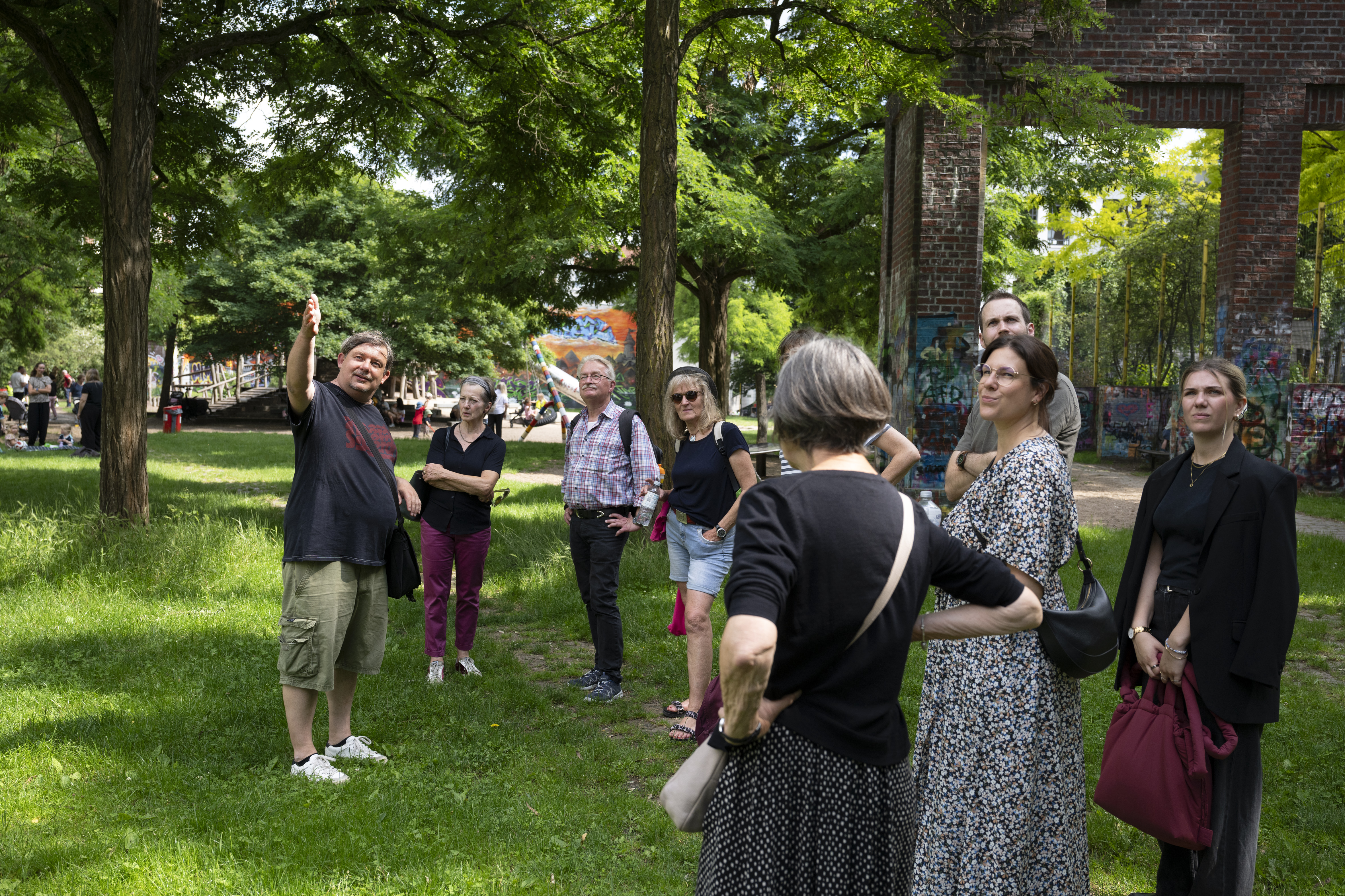 Eine Gruppe von Menschen steht auf einer Wiese in einem Park im charmanten Eppendorfer Viertel und lauscht einem Stadtführer, der auf verschiedene Orte zeigt und über die Geschichte von Hamburg-Hoheluft berichtet.
