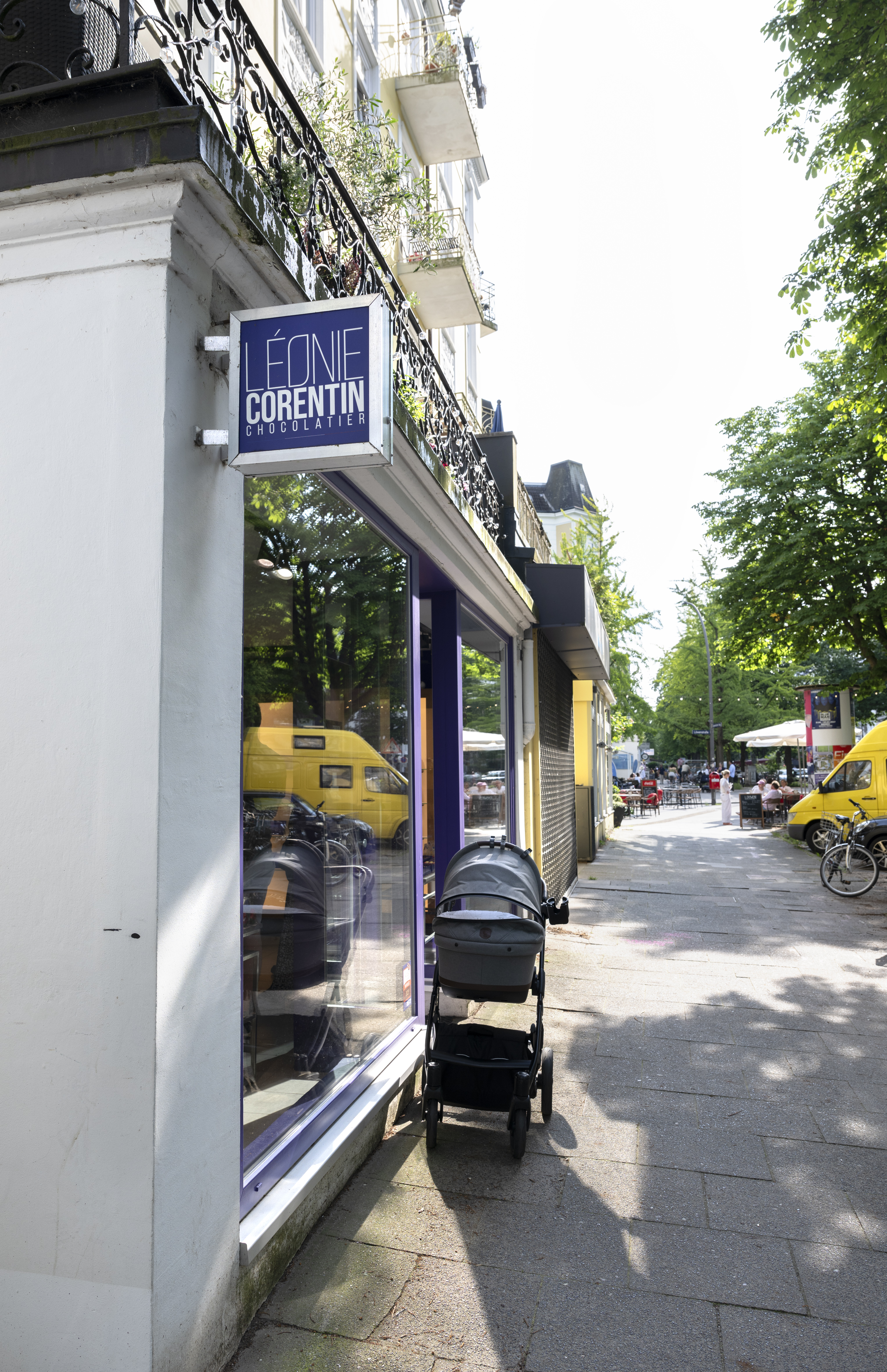 Ein Kinderwagen steht vor einem Geschäft mit dem Schild „Léonie Corentin“ in einer sonnigen, von Bäumen gesäumten Straße in Hamburg-Hoheluft, einem charmanten Viertel, das für seine einladende Atmosphäre bekannt ist.