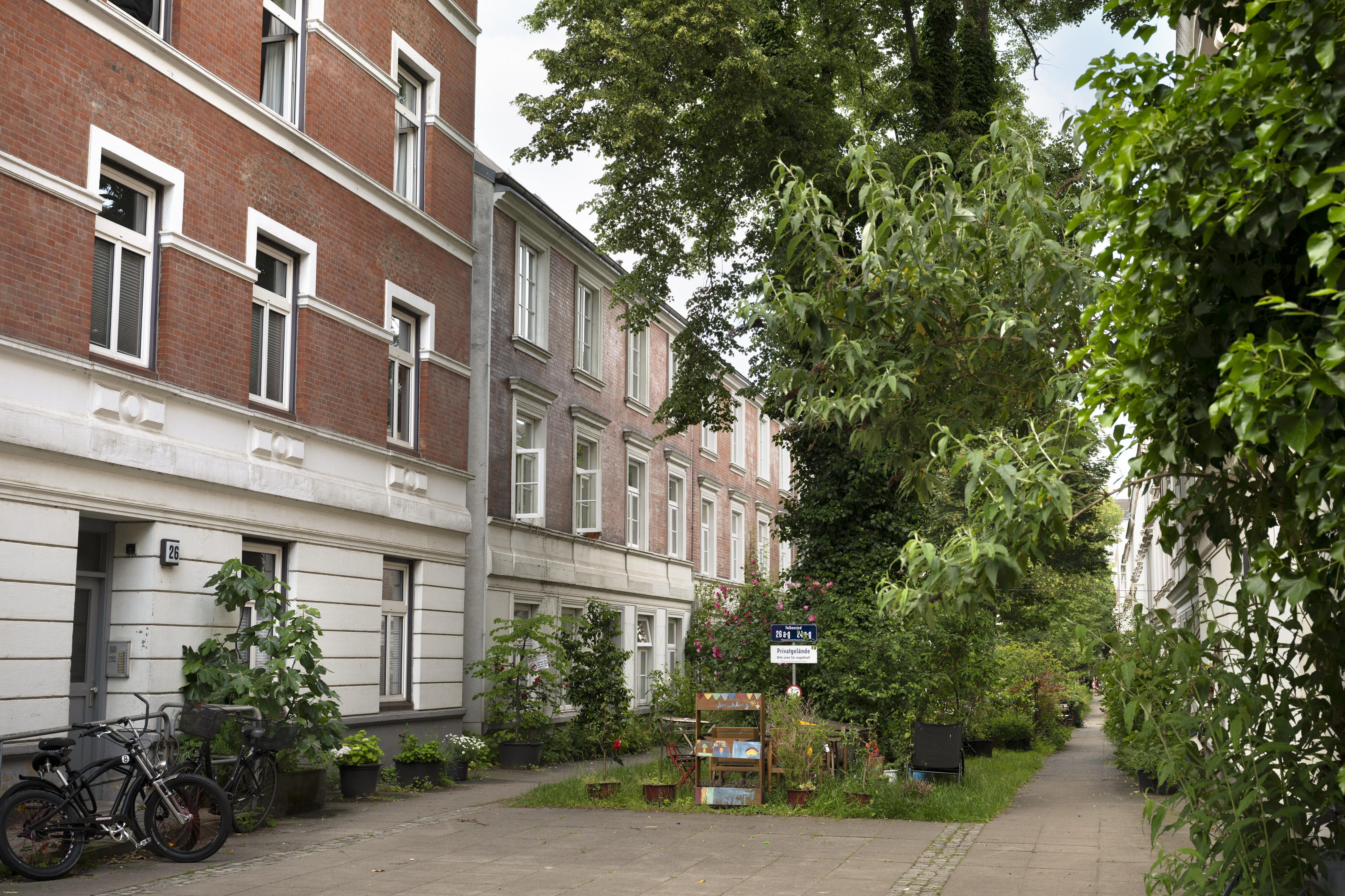 Eine ruhige Wohnstraße in Hamburg-Hoheluft mit Topfpflanzen, Fahrrädern und einer kleinen Holzbank vor den Wohnhäusern – das fängt das „Charmantes-Viertel“-Flair dieses reizenden Viertels perfekt ein.