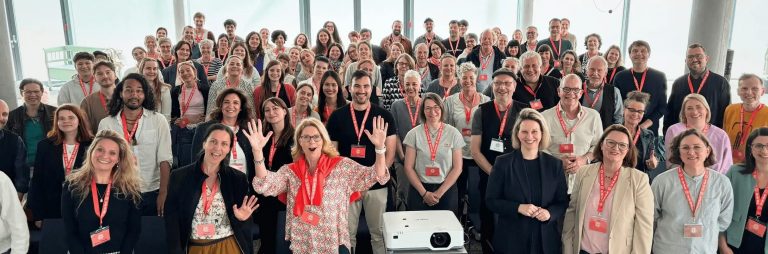 Eine große Gruppe lächelnder Menschen mit roten Schlüsselbändern, die gemeinsam in einem Innenraum vor großen Fenstern posieren und eine lebendige Teamkultur sowie eine gemeinsame Leidenschaft für die Karriere widerspiegeln.