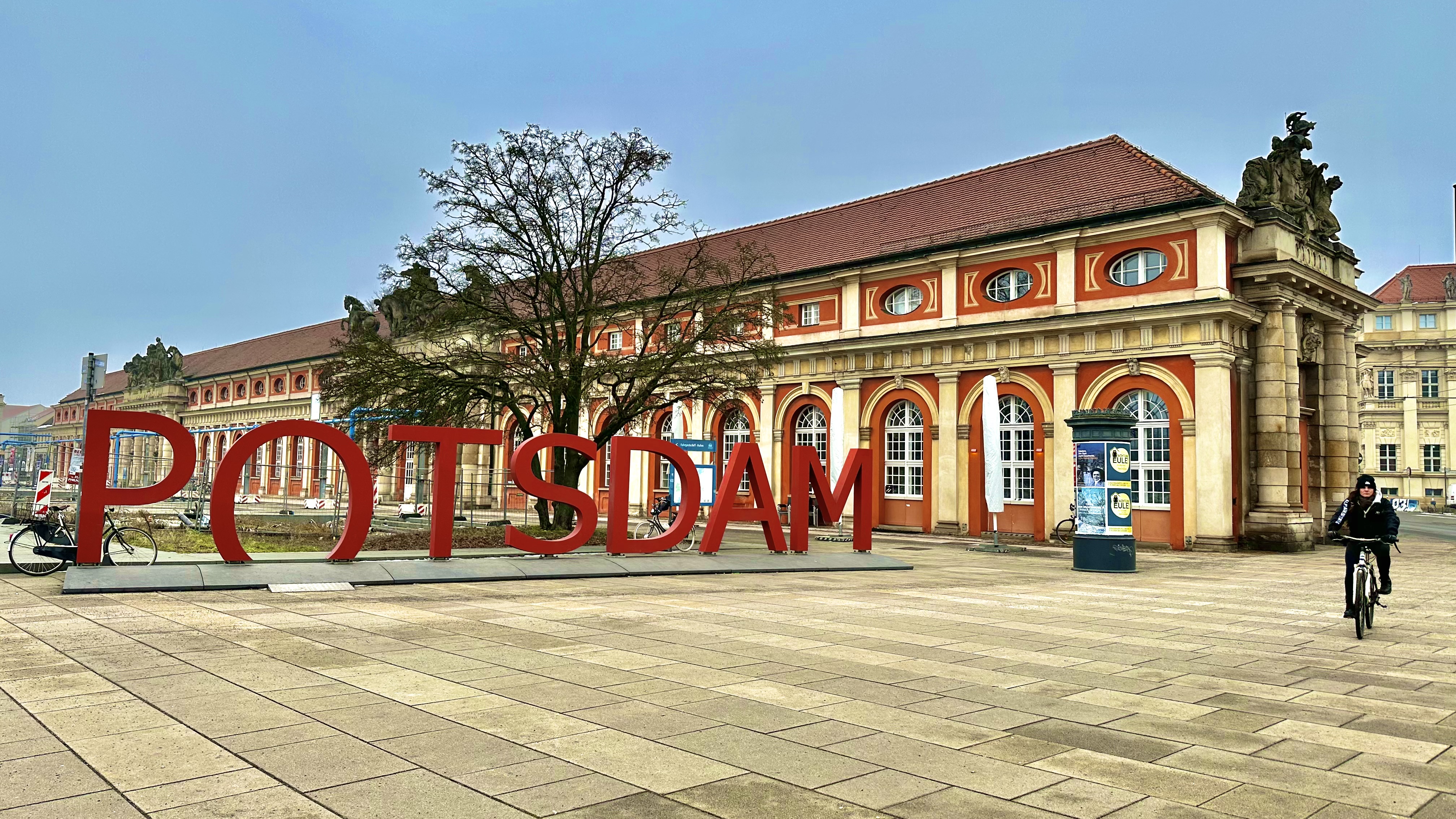 Ein großes rotes „POTSDAM“-Schild vor einem historischen Gebäude mit Bogenfenstern – perfekt für Ihren Tagesausflug nach Potsdam –, während ein Radfahrer vorbeifährt.