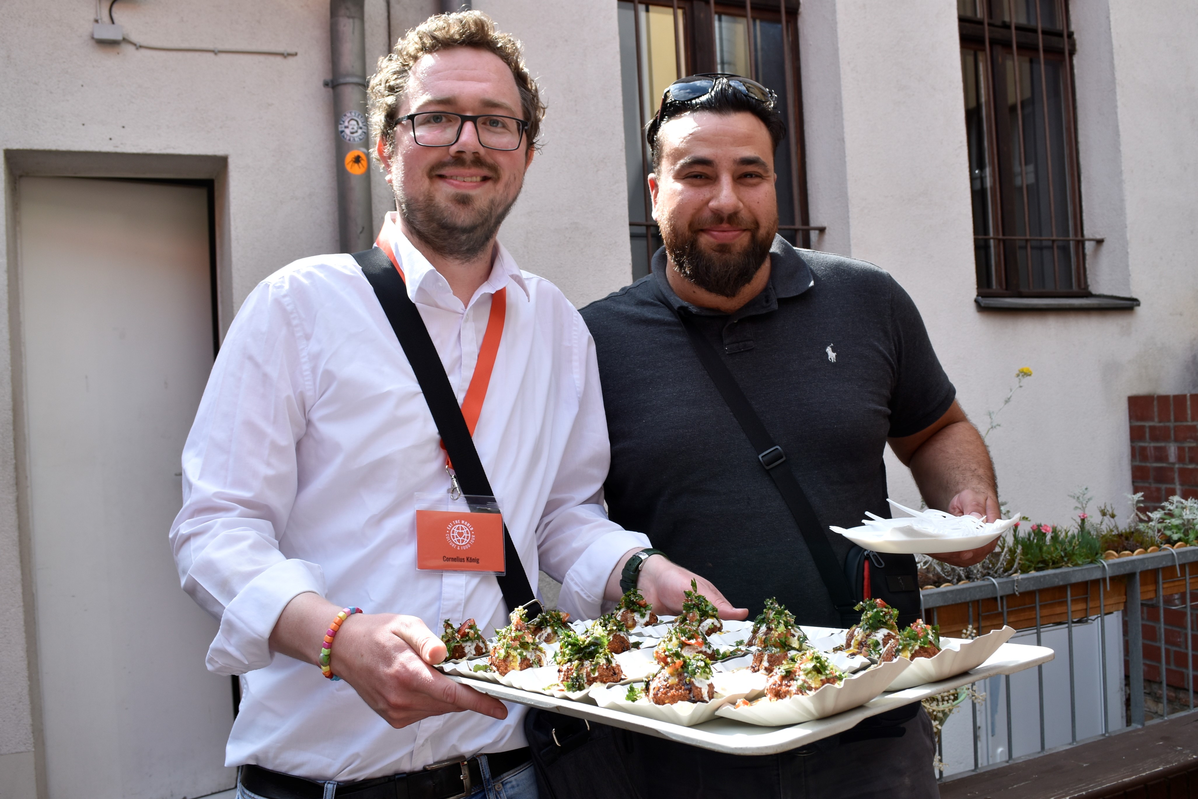 Zwei Männer, die während eines Tagesausflugs in Potsdam im Freien lächelnd Tabletts mit Essen in den Händen halten; einer trägt ein Abzeichen, beide sind leger gekleidet.